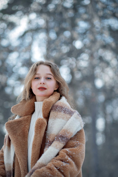 一个戴着针织帽、戴着长围巾、穿着毛皮大衣的金发女孩微笑着穿过森林。女人戴着白色手套抓雪。冬天在街上。平安夜在公园。欧洲长相的女孩享受生活。新年的心情，礼物，s图片下载