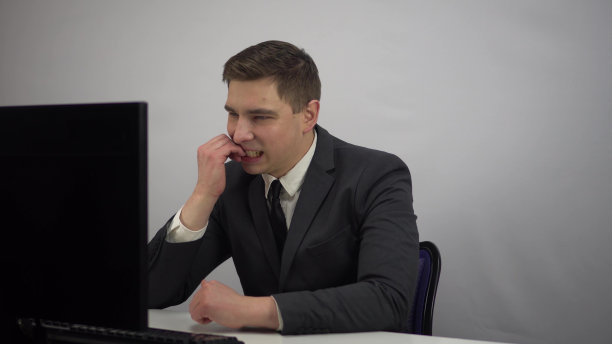 一位年轻的商人在工作场所紧张地咬指甲。穿着西装的IT专家坐在办公室里看着电脑显示器，神情紧张。图片下载