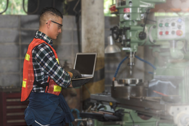 在模糊的工厂背景下，工程师一边用笔记本电脑监控工作，一边摆弄金属车床。图片下载