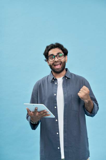 兴奋的阿拉伯男人手持平板电脑庆祝胜利，蓝色背景图片下载