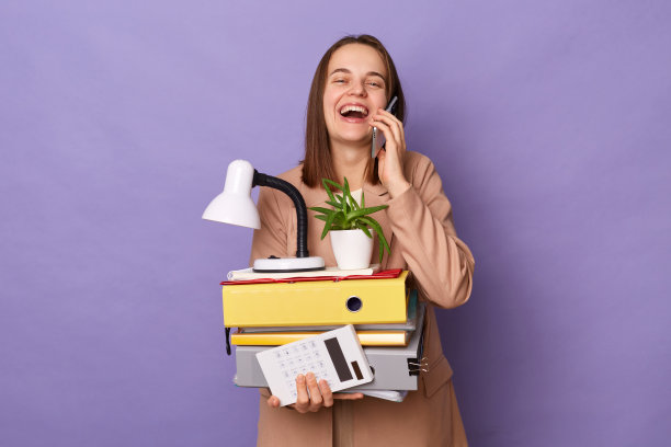 照片中，穿着米色夹克的快乐女子笑着拿着很多文件文件夹，在淡紫色的背景下隔离，打电话，吹嘘她的新工作，高兴。图片下载