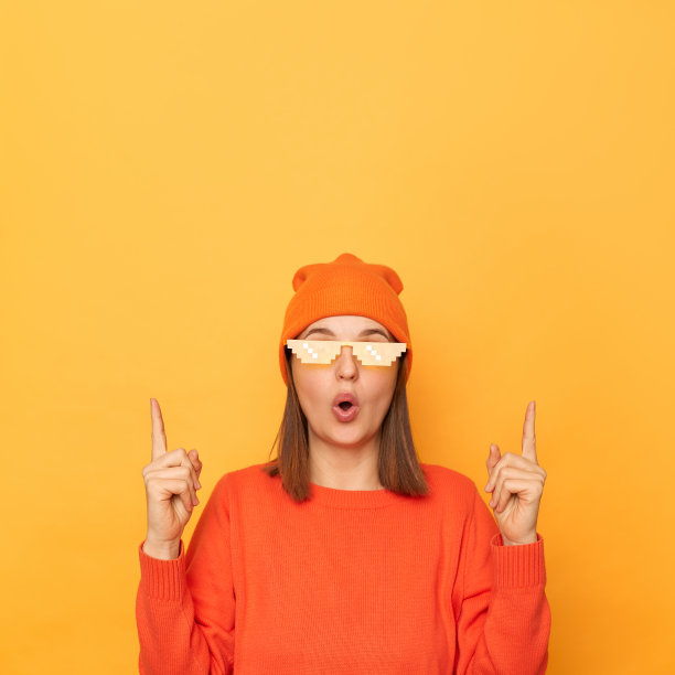 照片中的年轻女子穿着橙色套头衫，戴着我的世界风格的无檐帽和眼镜，指着广告的复制空间，孤立地站在黄色背景上。图片下载