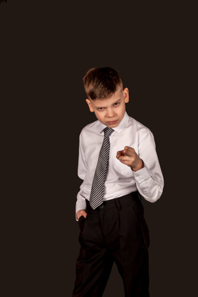 坏脾气的男孩皱着眉头用手指指着镜头，穿着白色衬衫和领带，黑色孤立的背景，看着。愤怒的孩子在工作室摆姿势。童年情感概念图片下载