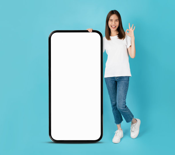 美丽的亚洲女子展示ok标志，站在空白屏幕的大型智能手机模型面前，在蓝色背景上微笑。图片下载