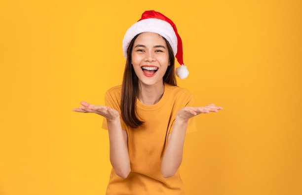 开朗美丽的亚洲女人戴着红色的圣诞帽，穿着橙色的t恤，站在橙色的背景上。图片下载
