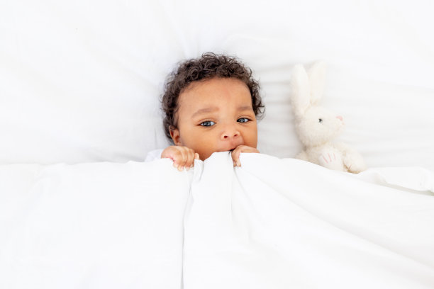 微笑的非裔美国小婴儿在白色的床上睡觉，毯子下有一个玩具图片下载