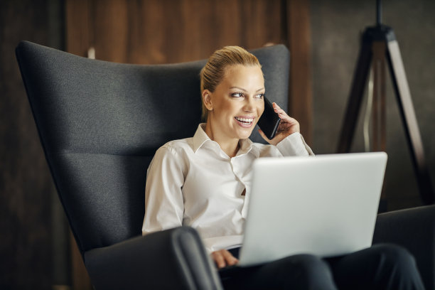 一位忙碌的女士出差期间坐在酒店房间里，坐在椅子上进行重要的电话交谈，膝上放着一台笔记本电脑。图片下载