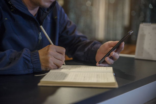 男性手里的计划员写着每天的约会，在日历簿上，在家里的工作空间。在办公桌上使用智能手机，在日记上记录计划和行程(假期旅行)。计划的概念。图片下载