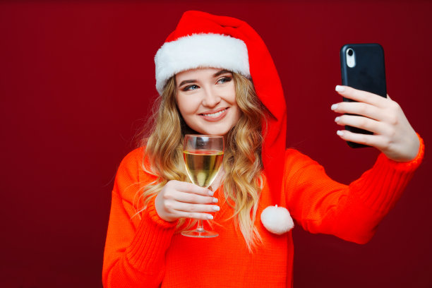 一个女人戴着圣诞老人的帽子，红色背景是香槟和智能手机。图片下载