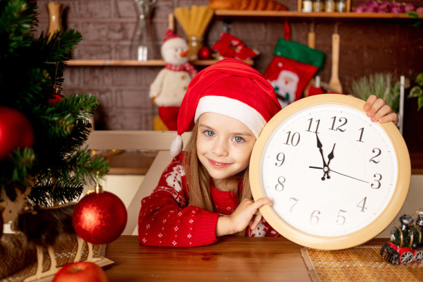 在黑暗的厨房里，一个戴着圣诞帽、拿着大钟的小女孩在一棵挂着红球的圣诞树旁快乐地微笑着，这是新年和圣诞节的概念图片下载