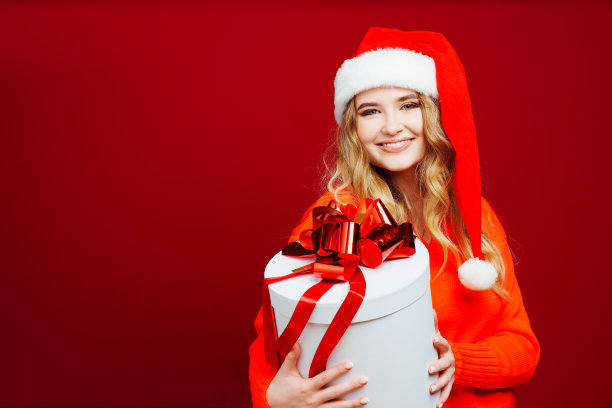 一个美丽的年轻女子，戴着圣诞老人的帽子，红色背景上有一份礼物。图片下载