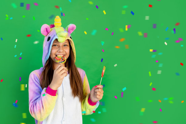 一个快乐的小女孩穿着独角兽服装，在绿色的孤立背景上有一条蛇，吃着大棒棒糖。文本空格。节日的概念图片下载