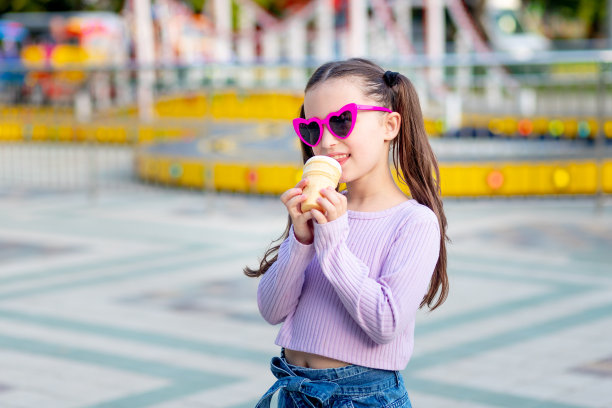 夏天，一个小女孩在游乐园的旋转木马旁吃着冰淇淋，戴着太阳镜快乐地笑着，周末和学校假期的概念图片下载