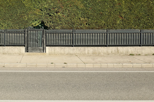 房子的栅栏由一个高的篱笆和一个小的门，金属栏杆和一个低的混凝土墙组成。前方是水泥人行道和柏油路。图片下载