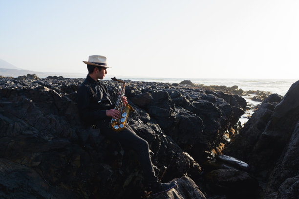 年轻的拉丁萨克斯演奏家坐在海滩上的岩石上放松地看着日落在地平线上的灵感时刻图片下载