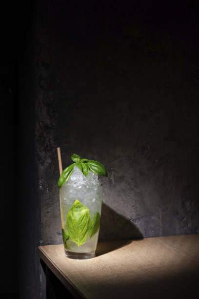 一杯鸡尾酒，加了冰块、罗勒和一根吸管，杯子放在桌子上，背景是一堵石墙图片下载