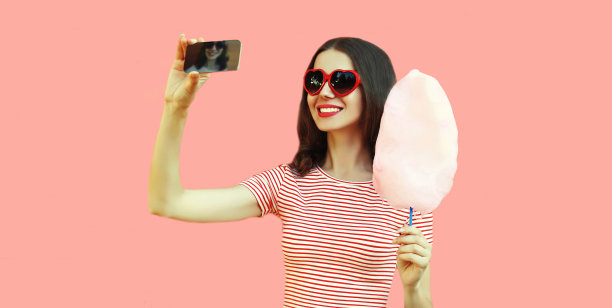 快乐的微笑年轻女子的肖像与棉花糖自拍与智能手机戴红色心形太阳镜在粉红色的背景图片下载