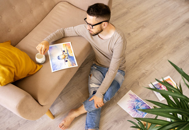 艺术家正坐在地板上，端着一杯咖啡。看着一幅有飞机的画，很伤心，梦想着在大流行期间乘飞机度假。真实情感生活方式图片下载
