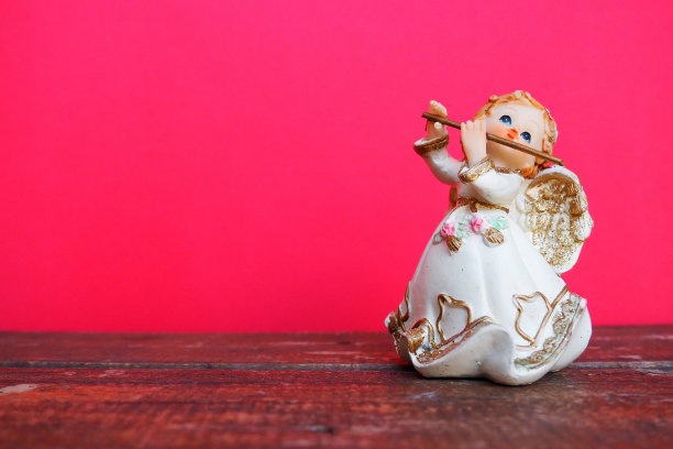 守护天使吹奏笛子。一个红发女孩的雕像，穿着白裙子，带着翅膀和烟斗。新年或圣诞假期背景。副本的空间。粉色木制背景。文本的空闲空间。图片下载