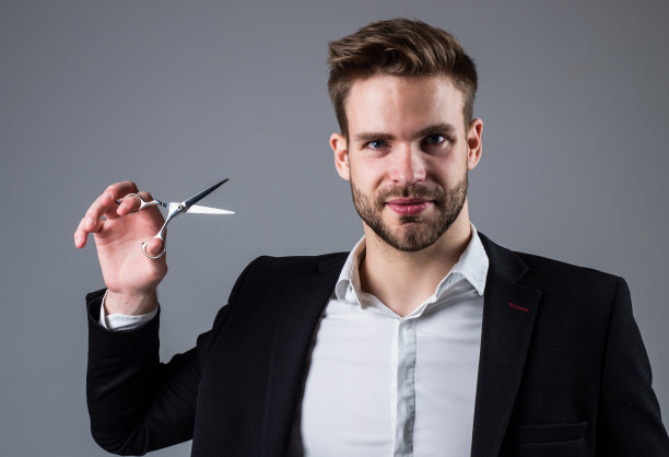 时尚是他的生命。男士发型设计和修饰。穿着西装的年轻帅哥拿着剪刀。男理发师与工具。留着大胡子的商人。时尚与美丽。理发店的概念图片下载