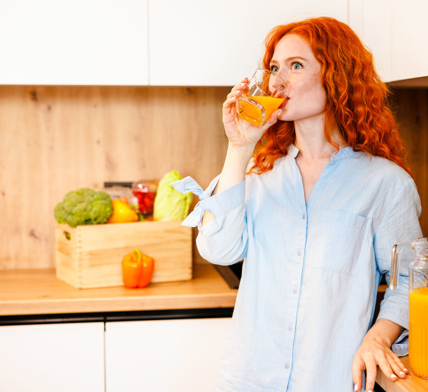 漂亮的年轻女子一边看一边在家里的厨房里喝果汁。图片下载