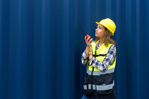 年轻的白人妇女，穿着安全背心和黄色安全帽，拿着对讲机，站在一个大型货物集装箱前。图片下载