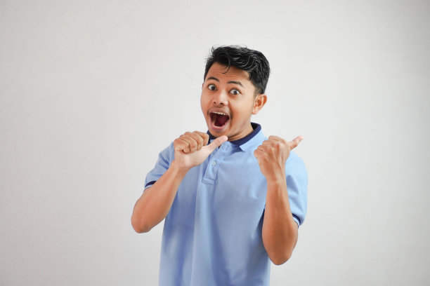 震惊的亚洲男子张开嘴，手指指向后面穿着蓝色t恤孤立在白色背景上图片下载