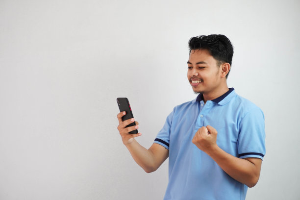兴奋或快乐的肖像年轻的亚洲男子握着手机，握紧的拳头，穿着蓝色的polo衫孤立在白色背景上图片下载