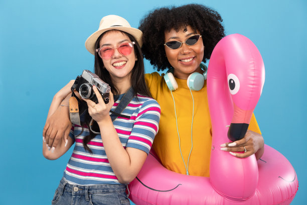 快乐的微笑非洲和亚洲妇女拿着火烈鸟橡胶环，享受他们的暑假。年轻的非洲裔美国妇女孤立在蓝色背景的假期与旅行。图片下载