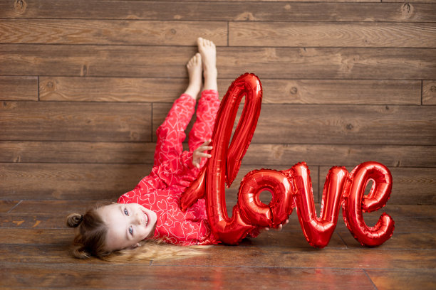 一个穿着红色睡衣，拿着一个写着“爱”字样的大气球的快乐的小女孩躺在地板上深棕色的木制背景上笑着，情人节的概念，一个文本的地方图片下载