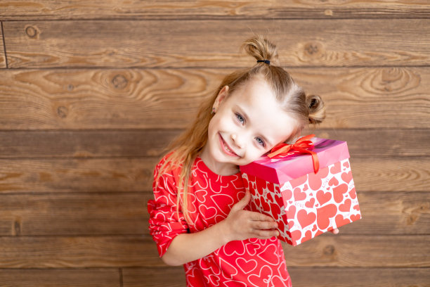一个穿着红色睡衣，手里拿着大礼物的小女孩在深棕色的木制背景上笑着笑着，情人节的概念，一个发短信的地方图片下载