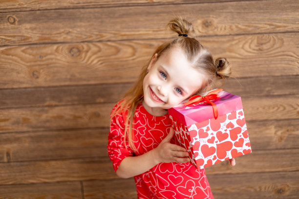 一个穿着红色睡衣，手里拿着大礼物的小女孩在深棕色的木制背景上笑着笑着，情人节的概念，一个发短信的地方图片下载