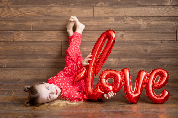 一个穿着红色睡衣，拿着一个写着“爱”字样的大气球的快乐的小女孩躺在地板上深棕色的木制背景上笑着，情人节的概念，一个文本的地方图片下载