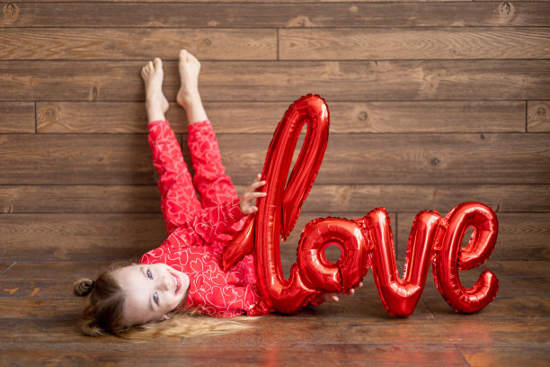 一个穿着红色睡衣，拿着一个写着“爱”字样的大气球的快乐的小女孩躺在地板上深棕色的木制背景上笑着，情人节的概念，一个文本的地方图片下载