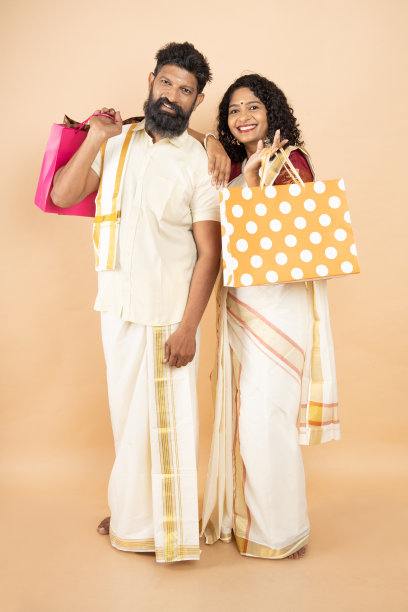 快乐的南印度夫妇穿着传统服装，拿着购物袋，在米色背景上一起庆祝节日。节日促销，复印空间。图片下载