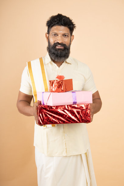 快乐的南印度男子穿着传统的白色连衣裙，手持礼盒孤立在米色背景的肖像。节日的庆祝活动。图片下载