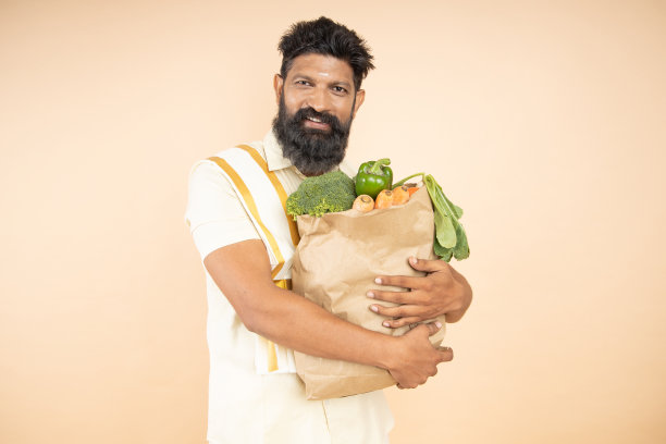 快乐的南印度男子拿着杂货店纸袋，米色背景上有食物。影棚拍摄。图片下载