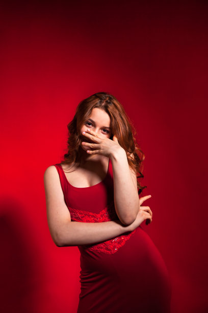 肖像笑少年封面女孩卷发在时尚的红色连衣裙覆盖脸与手在红色背景，看着相机。黑发女士在工作室摆姿势。时尚风格概念。本空间图片下载