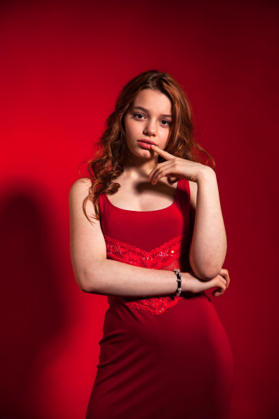 时髦的十几岁封面女孩卷发摆造型在红色背景的时尚红色连衣裙，看着相机。年轻漂亮的黑发女士在工作室。时尚风格概念。复制广告的文本空间图片下载