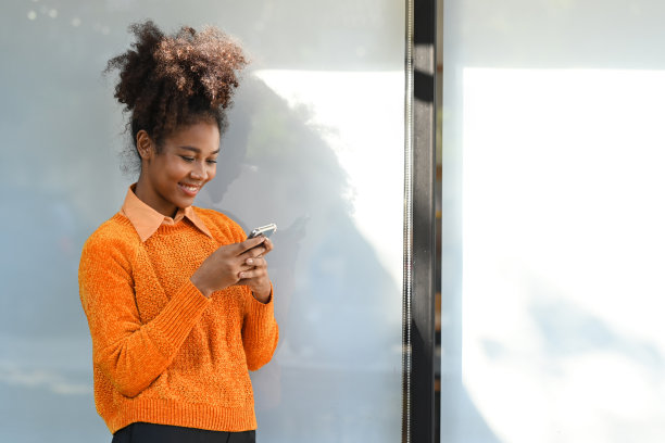 愉快的年轻女子阅读短信智能手机，而站在办公大楼外的城市。图片下载