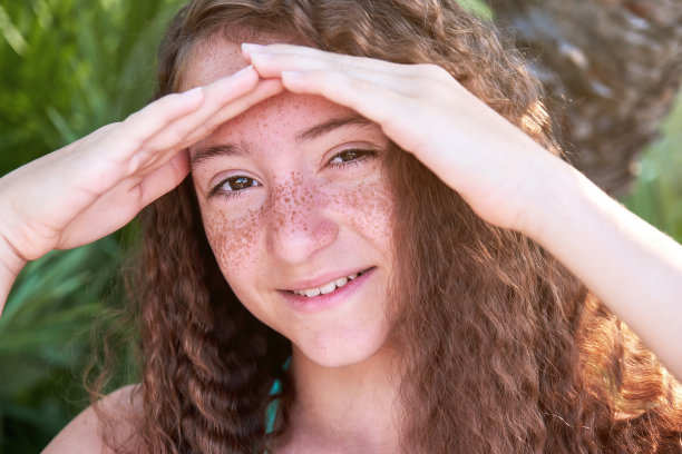 一个有雀斑的卷发少女眯着眼睛，用手遮着太阳。图片下载