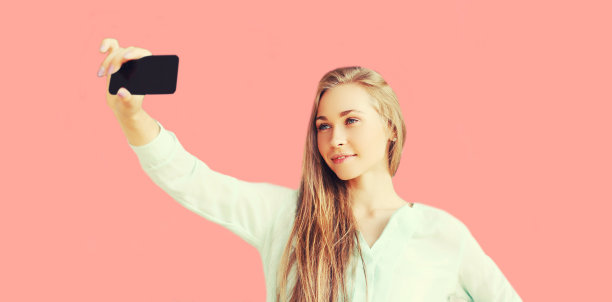 快乐的金发微笑的女人在粉红色背景上用智能手机自拍的肖像图片下载