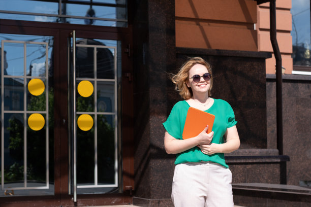 一个快乐的女研究生，戴着墨镜，拿着平板电脑，书本，笔记本靠在大学大楼上。图片下载