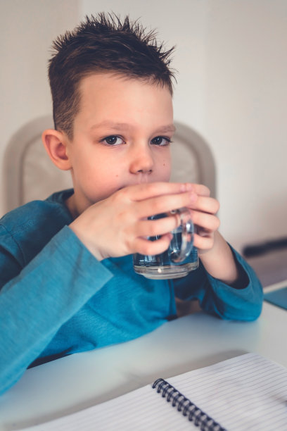 健康的孩子边做作业边喝水，开朗的学生坐在电脑前的桌子前，在线观看视频课程，家庭内部，复制空间。图片下载