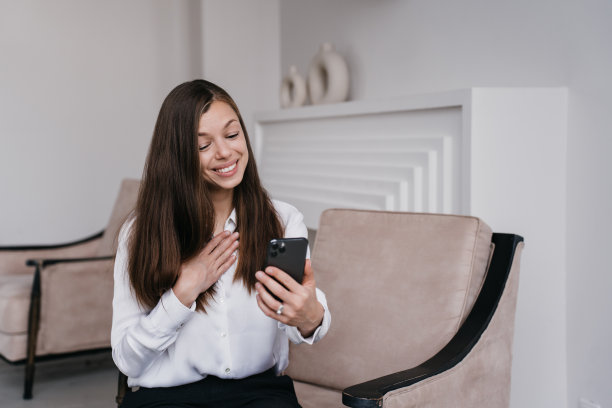 感激的意大利黑发年轻女子视频通话使用电话坐在椅子上，笑容灿烂。女学生在网上和朋友聊天时捂着胸口表示感谢。成功的女性。图片下载