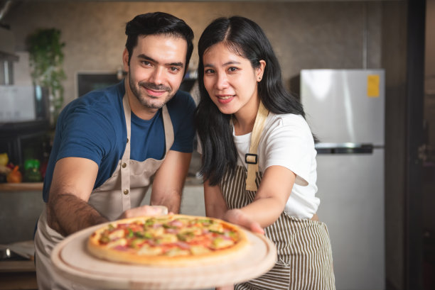 年轻夫妇家庭白人男子和亚洲妇女穿着围裙享受在家里厨房做披萨。小型商业餐厅和美食。图片下载
