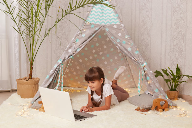 黑头发扎着小辫子的小女孩躺在棚屋里，拿着笔记本电脑，在网上看卡通片或有趣的视频，在她舒适的房间里度过周末。图片下载