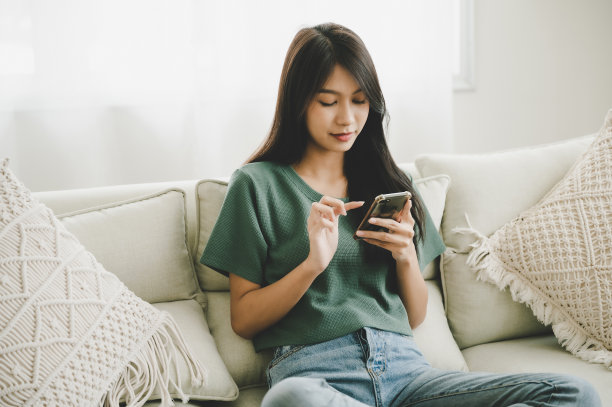 快乐的年轻亚洲女子在家里放松，她坐在沙发上，用手机图片下载
