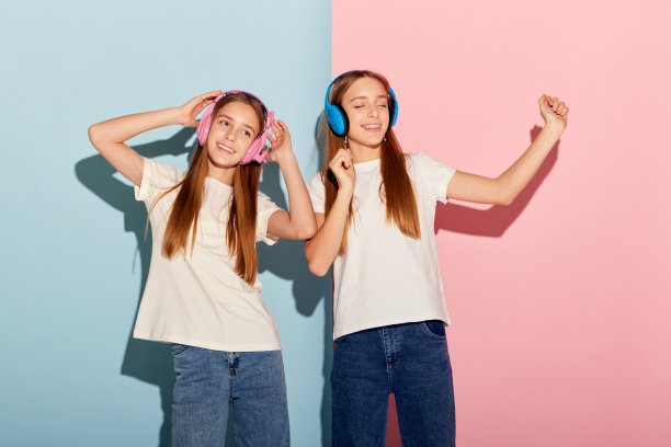 音乐。快乐的年轻女孩们，两姐妹朋友戴着彩色耳机，隔着粉蓝色的工作室背景。时尚、关系、情感、美丽和青春图片下载