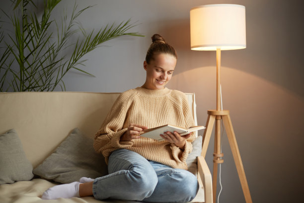 照片的白人年轻成年有吸引力的女子穿着米色套头衫和牛仔裤坐在咳嗽和读书，微笑，有愉快的面部表情，对小说的情节感兴趣。图片下载
